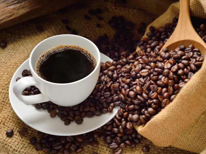 Taza de café negro recién preparado sobre una mesa de madera con granos de café esparcidos